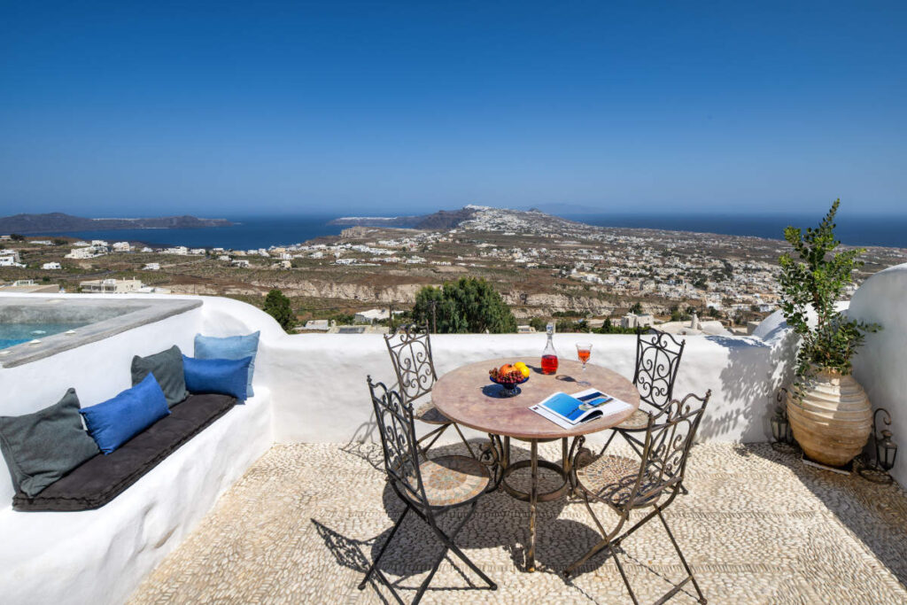 The outdoor patio at Villa Santa Croce features a sleek metal table, chic chairs, and a captivating book. Refreshing drinks and vibrant fruit sit atop, perfectly complementing the breathtaking sea and hilltop views under Santorini's clear blue sky.
