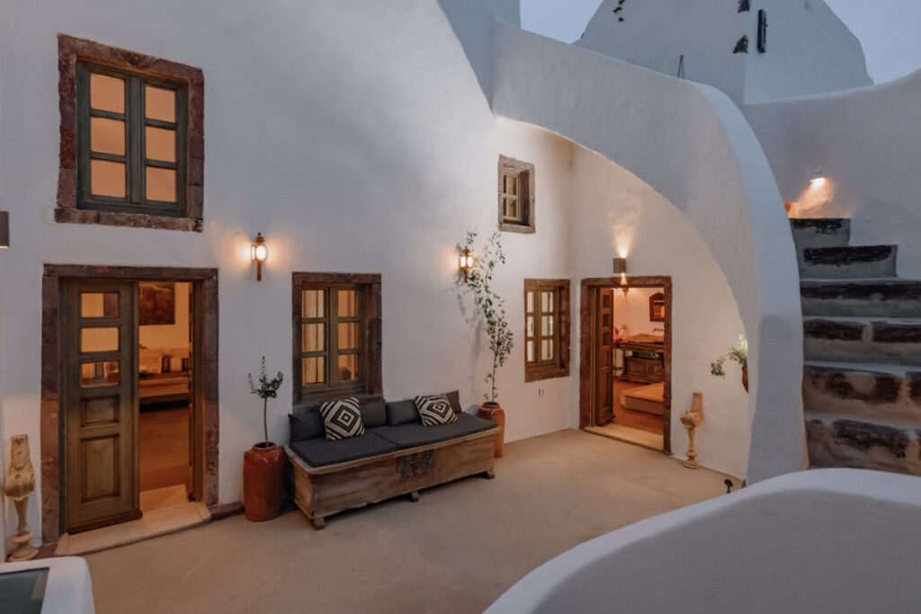 Discover the charm of Villa Santa Croce's outdoor courtyard, featuring whitewashed walls, rustic wooden doors, and steps leading up. Relax on a cushioned bench beneath warm wall lamps and enjoy the ambience enriched by decorative plants.