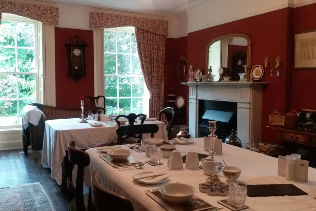 In the vintage dining room of Dromore House, red walls complement a cozy fireplace and large windows. Tables set with white tablecloths sparkle with fine dishes and silverware, while antique clocks and decor adorn the walls and mantel.