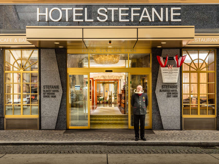 A doorman in uniform stands at the entrance of Hotel Stefanie Wien, a 4 star hotel Vienna, with elegant glass doors and gold accents welcoming guests to this historic building.
