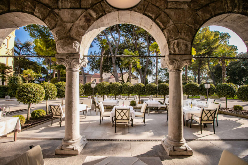 Outdoor patio seating at Hotel Sveti Jakov Opatija, with tables and chairs surrounded by green shrubbery and trees, framed by stone arches.