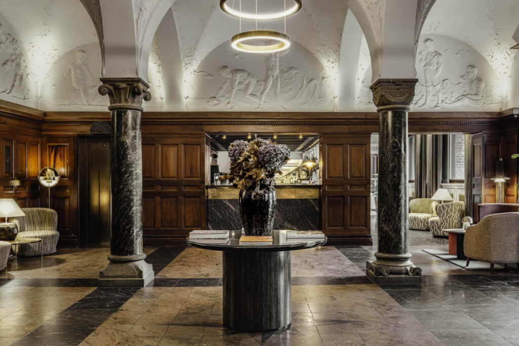 The elegant Hotel Ascot lobby in Copenhagen features a marble table adorned with flowers, an ornate ceiling, and wooden wall panels. Two columns and inviting seating areas complete this sophisticated space.