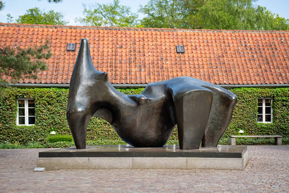 A large abstract bronze sculpture on a stone platform is displayed in a courtyard with cobblestone ground, ivy-covered wall, and red-tiled roof in the background.