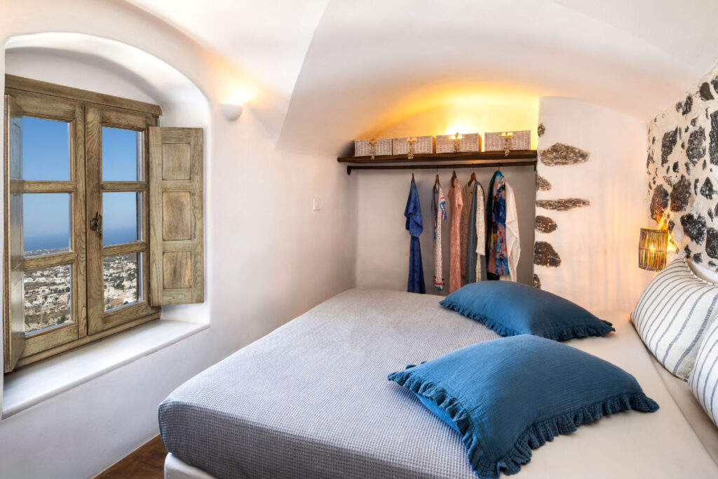 A cozy bedroom at Villa Santa Croce features a bed with blue pillows, a window offering a scenic view, and a wardrobe with several hanging clothes illuminated by ambient lighting. The walls boast rustic wooden and stone accents, embodying the serene charm of Santa Croce Villa in Santorini.