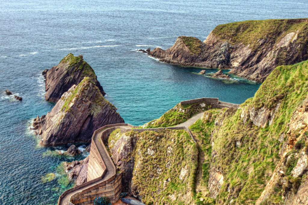 Aerial view of Dunquin Pier on coastal cliffs overlooking turquoise waters, with prominent rocky formations jutting into the sea.
