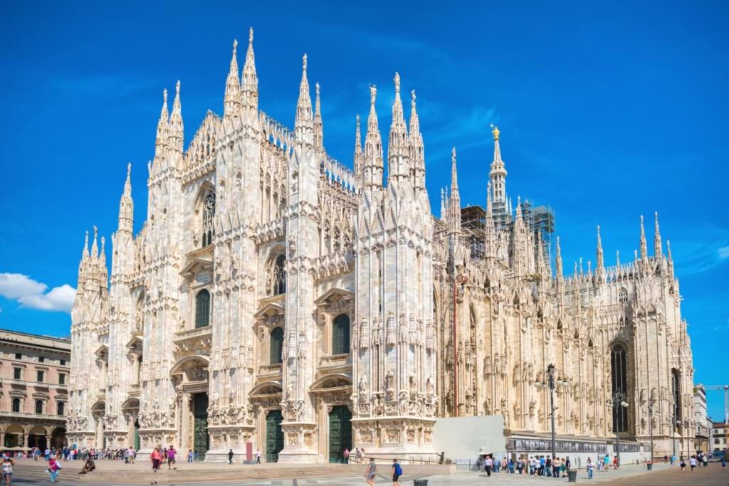 This Gothic-style cathedral, featured in the Milan Travel Guide, boasts intricate spires and statues. Visitors stroll across the plaza out front under a bright blue sky.