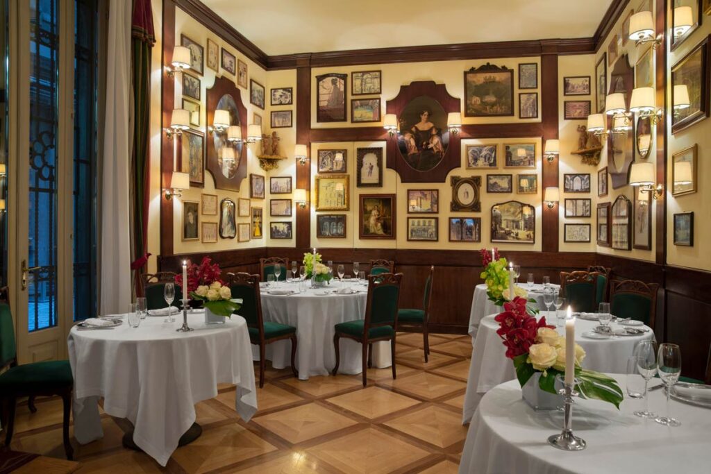 Elegant restaurant interior with round tables draped in white tablecloths and floral centerpieces, nestled in a space reflecting Milan history through walls adorned with numerous framed pictures and paintings.