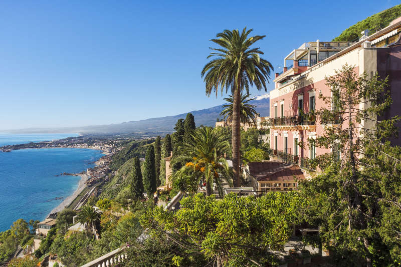 Coastal view of a Mediterranean town with lush greenery, a pink building, and palm trees near the renowned Hotel Villa Schuler in Taormina, Sicily. The distant shoreline sparkles under a clear blue sky.