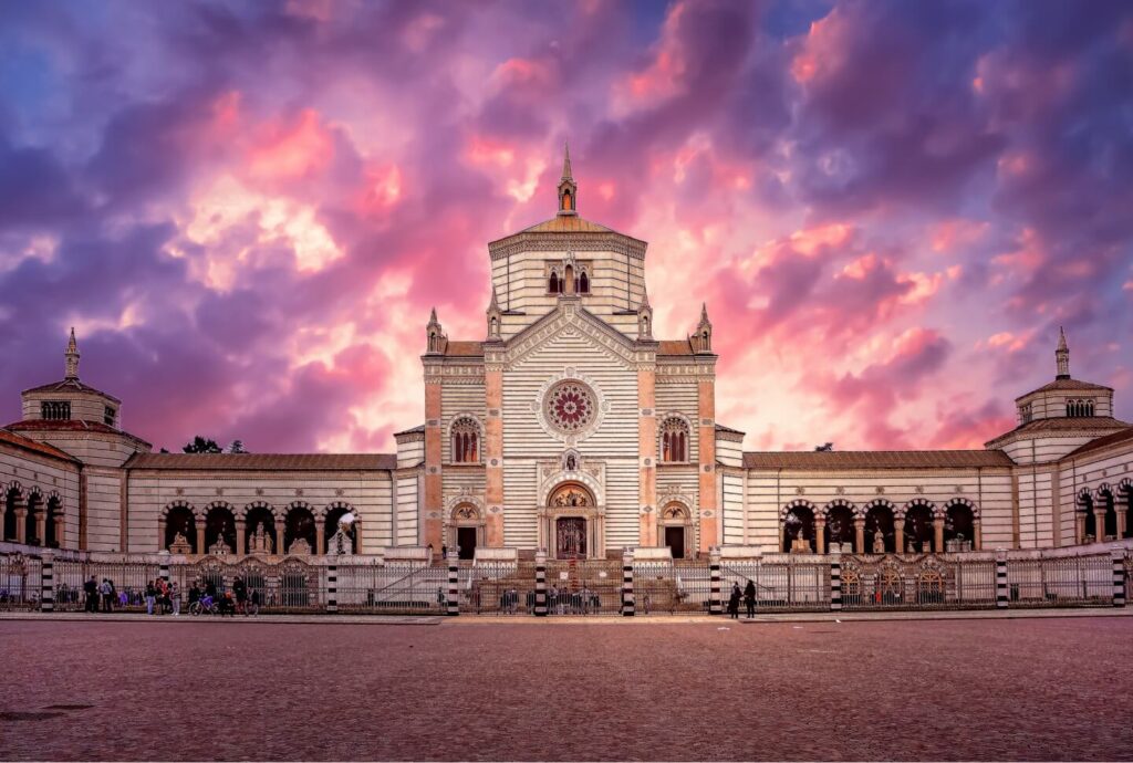The image captures a grand, striped building with ornate details under a vibrant, cloudy sky at sunset, illustrating a must-see in any Milan travel guide. The structure's arches and central tower stand majestically, while people and trees animate the background.