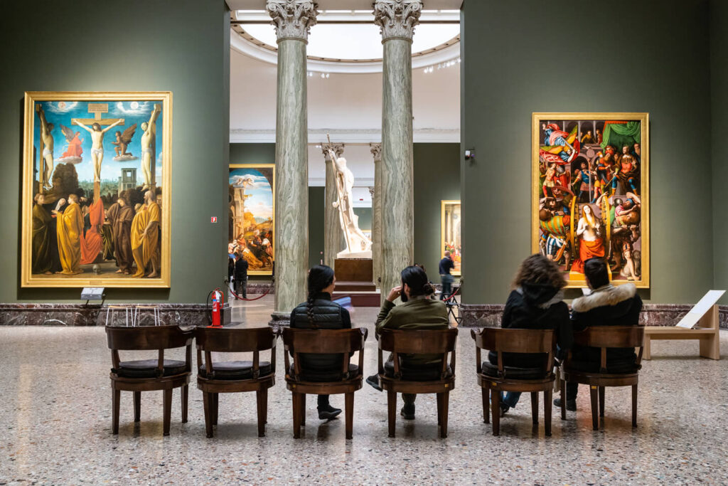 This Milan travel guide highlights a museum gallery with green walls, showcasing large framed paintings and a central statue. Five visitors are seated on wooden chairs, thoughtfully gazing at the artworks, immersing themselves in the rich cultural atmosphere.