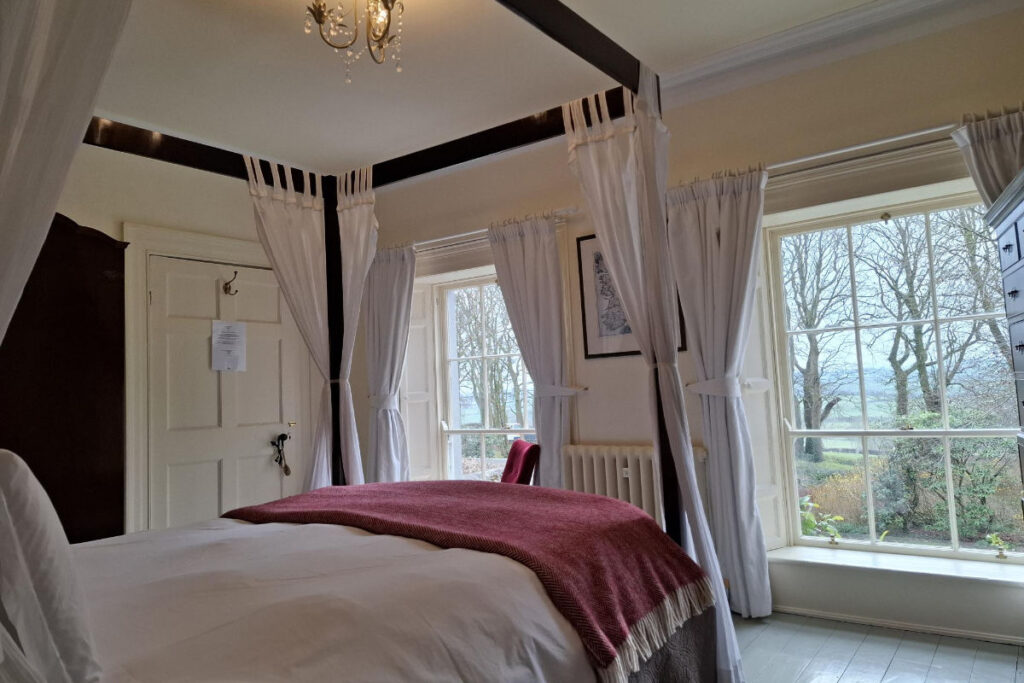 A cozy bedroom in Dromore House features a four-poster bed adorned with white curtains and a red blanket, while large windows offer a charming view of a garden filled with trees.