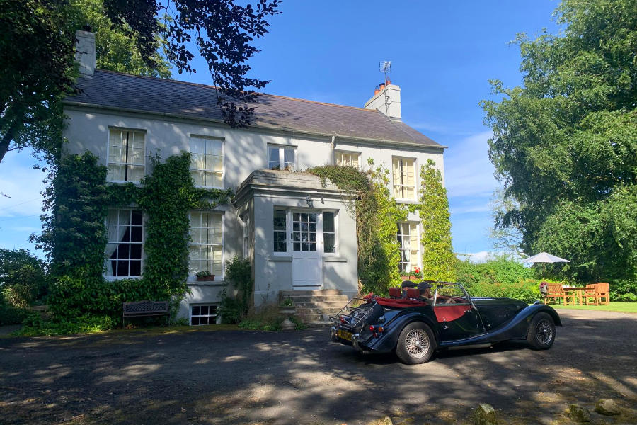 A vintage convertible car is parked in front of Dromore House, a large, ivy-covered two-story residence in Northern Ireland, basking in the sunshine.