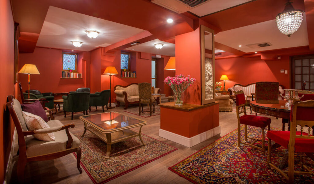 The warmly lit lounge at Hotel Polski features red walls, various seating arrangements, Persian rugs, and a decorative vase of flowers on a table.