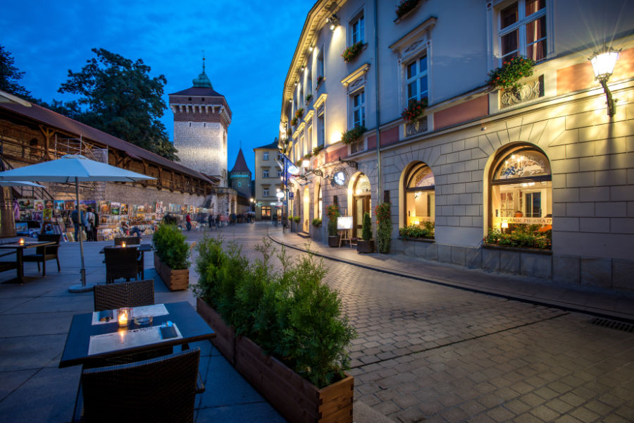 A cobblestone street lined with outdoor tables and potted plants leads to the charming Hotel Polski. Illuminated buildings and a historical tower stand proud under the twilight sky, offering a picturesque view for visitors.