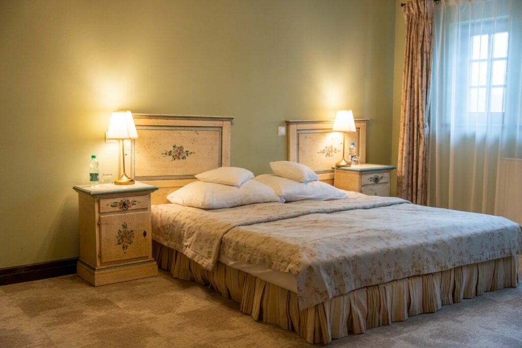 A bedroom with a double bed, two bedside tables with lamps, and floral decor reminiscent of Pałac Wojanów. A window with sheer curtains is on the right.