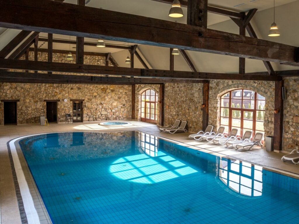 Indoor swimming pool with stone walls, wooden beams, a tiled floor, and lounge chairs reminiscent of Pałac Wojanów. Large arched windows allow natural light to illuminate the pool area.
