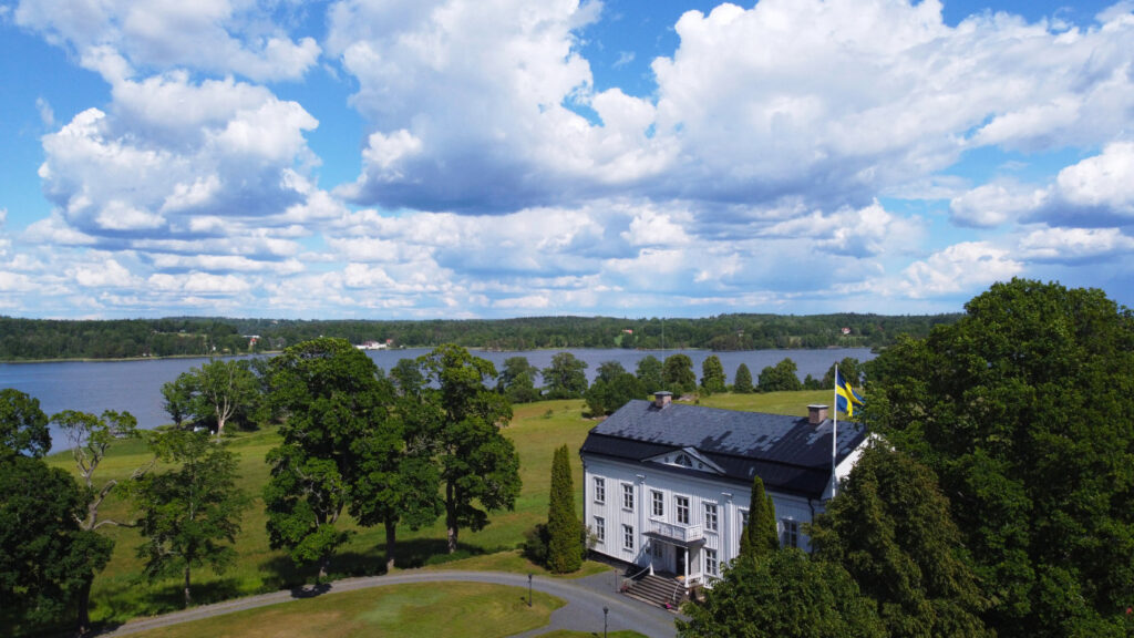 A large white building, Wallby Säteri, with a Swedish flag sits among trees near a river under a partly cloudy sky.