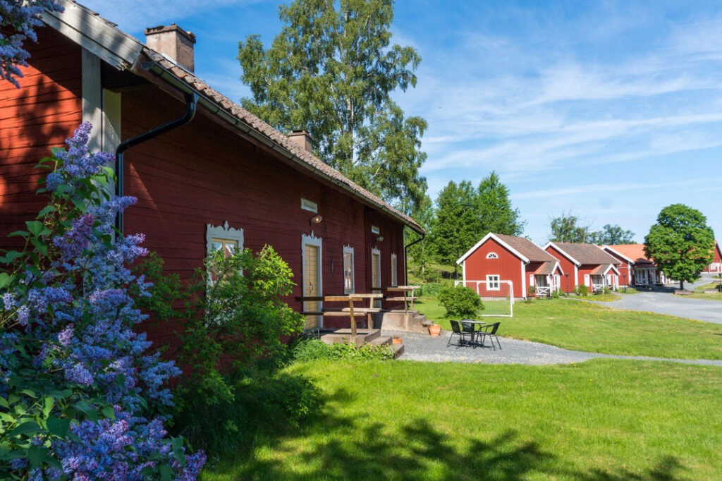 Red wooden houses with white trim at Wallby Säteri are set on a grassy area with outdoor furniture, trees, and lilac bushes under a clear blue sky.