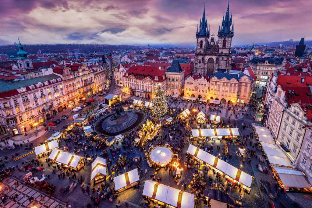 Aerial view of a bustling Christmas market in a historic city square, featuring a large church, colorful stalls, and a decorated tree at sunset.