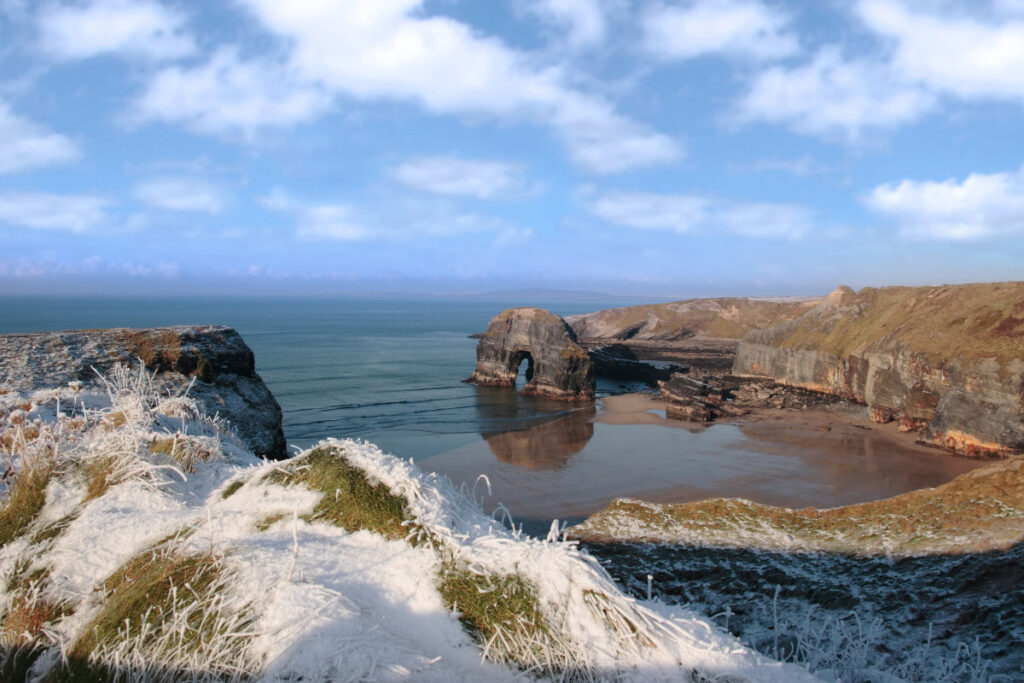 Coastal landscape with a large rock arch in the sea, surrounded by cliffs. Frost-covered grass graces the foreground under a blue sky with scattered clouds, creating an idyllic view reminiscent of scenic vistas by historic hotels to visit in winter.