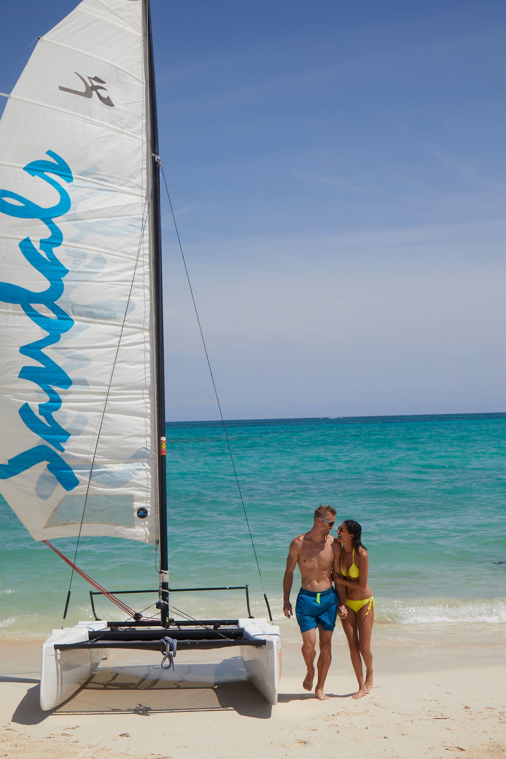 a couple laughing and walking away from the ocean near a sandals branded hobie cat