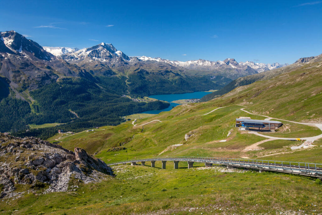 A scenic mountain landscape with a lake in the distance, winding roads perfect for European roadtrips, a wooden bridge, and a building on a grassy slope under a clear blue sky.