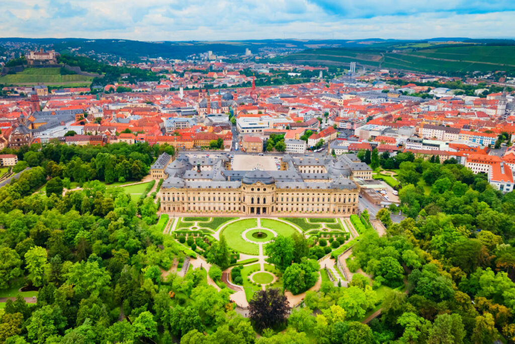 Aerial view of the large Würzburg Residence with expansive formal gardens, surrounded by a cityscape and green hills under a cloudy sky.