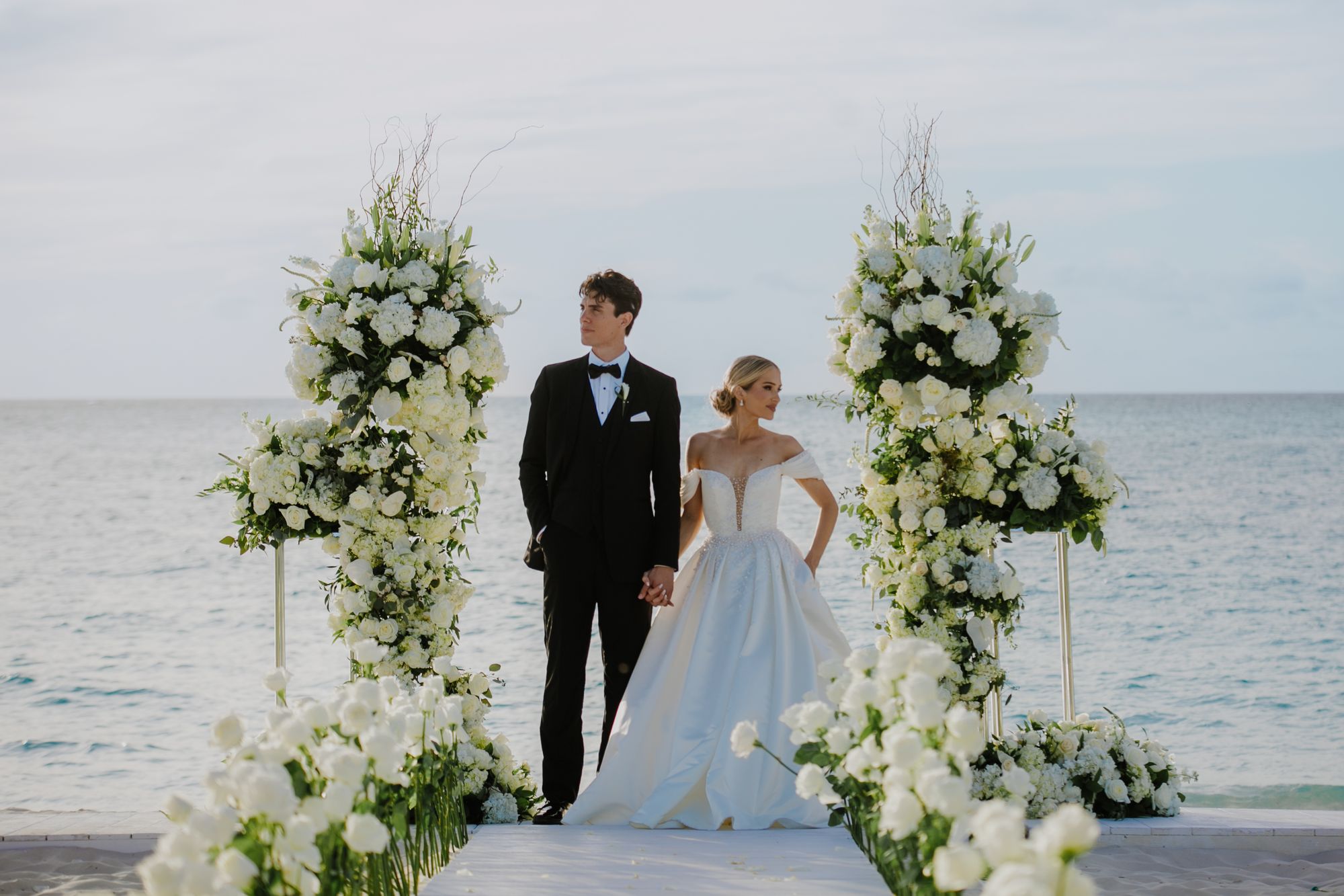 jackson and sarah holding hands looking in oposite directions standing in between white flowers and the beach in the background